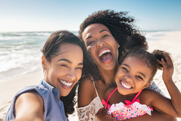 2 jeunes femmes souriantes en post-partum faisant un selfie avec leur petite fille sur une plage